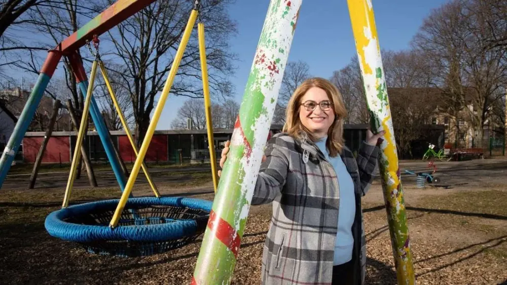 Der heutige Spielplatz an der Fritz-Tarnow-Straße: eine Frau steht zwischen zwei Pfosten mit abblätternder Farbe, dahinter eine blaue Rollstuhlschaukel.