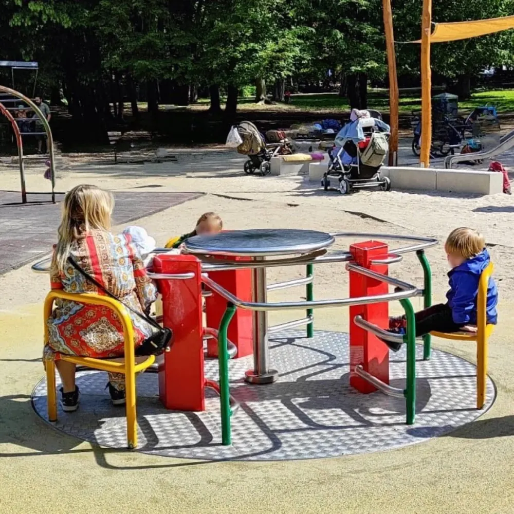 Eine Frau und zwei Kinder sitzen auf dem Rolli-Karussell im Günthersburgpark, dahinter der Sandspielbereich.