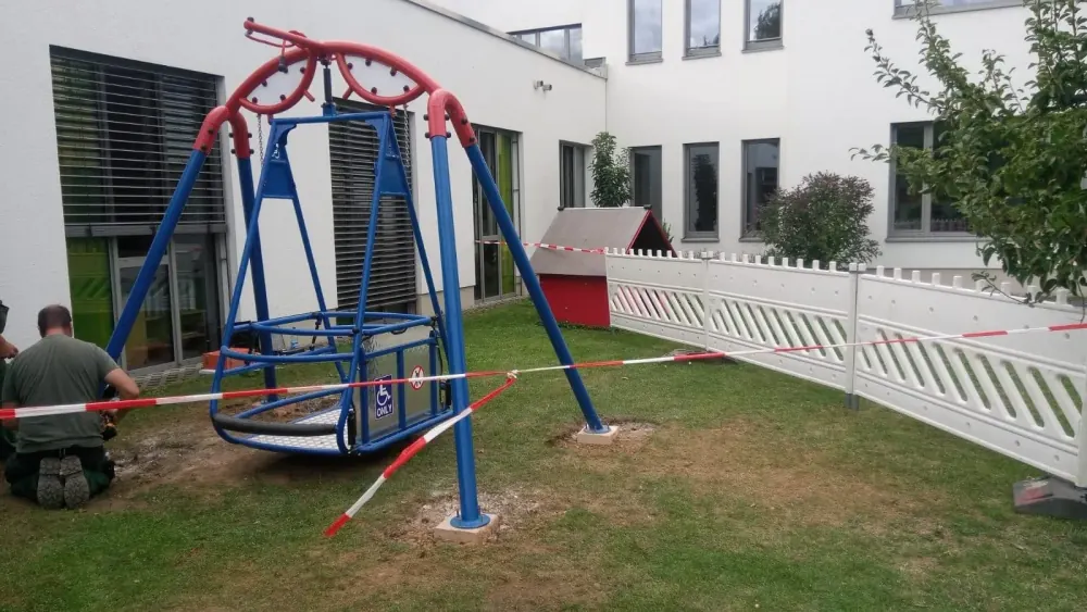 Die neu aufgebaute Rollstuhl-Schaukel auf dem Gelände der Martin-Buber-Schule, noch mit Absperrband gesichert. Ein Monteur arbeitet daneben.
